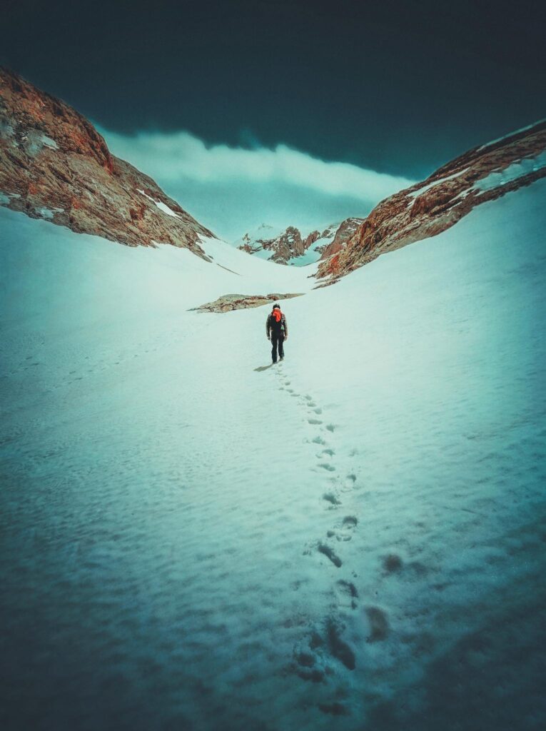 Back view of unrecognizable hiker in warm clothes walking on snowy mountain slope against blue sky