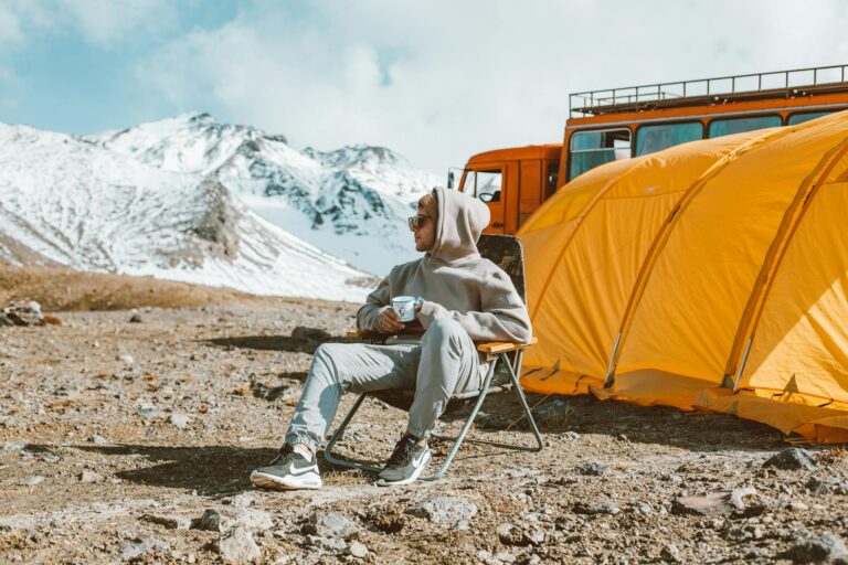 Full length of male camper sitting on folding chair next to tent in middle of mountain valley and enjoying views